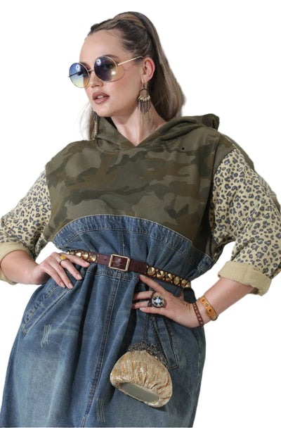 Woman wearing a green hooded top and blue denim dress with leopard print sleeves against a white wall.